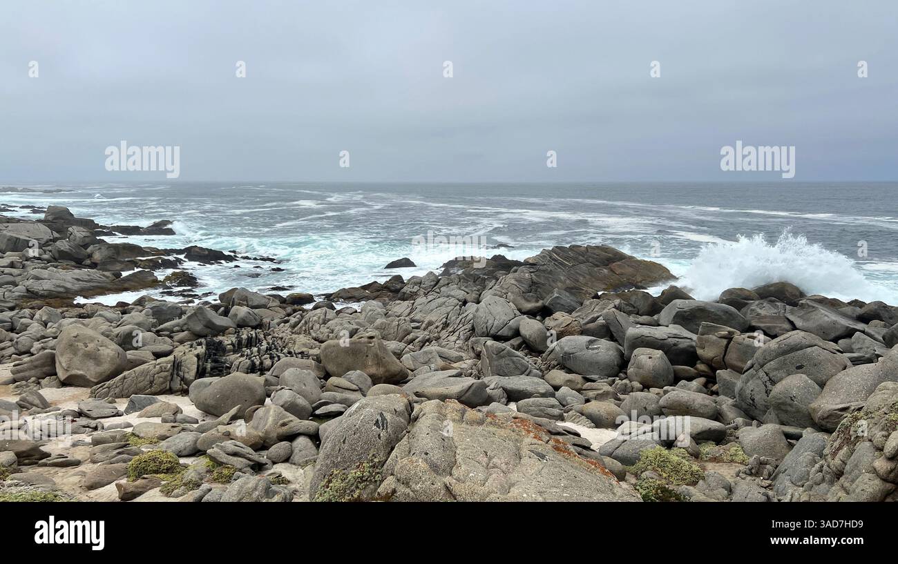 Felsige Küste mit Wellen, die unter bewölktem Himmel zusammenbrechen Stockfoto