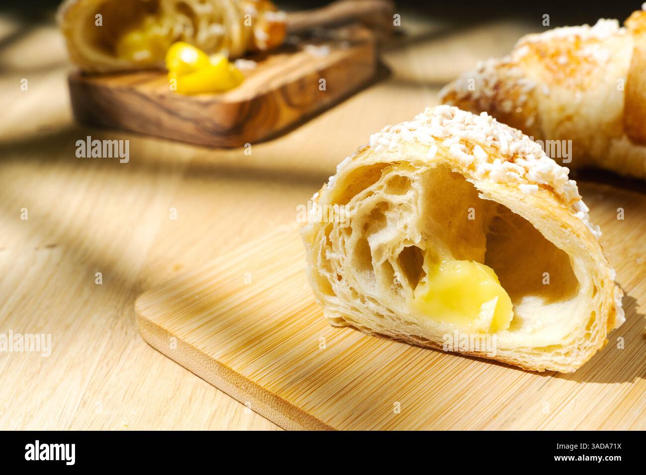 Nahaufnahme von geschnittenem Zitronencroissant auf einem hölzernen Schneidebrett. Leicht verschwommenes Croissant mit Zitronenschale im Hintergrund Stockfoto