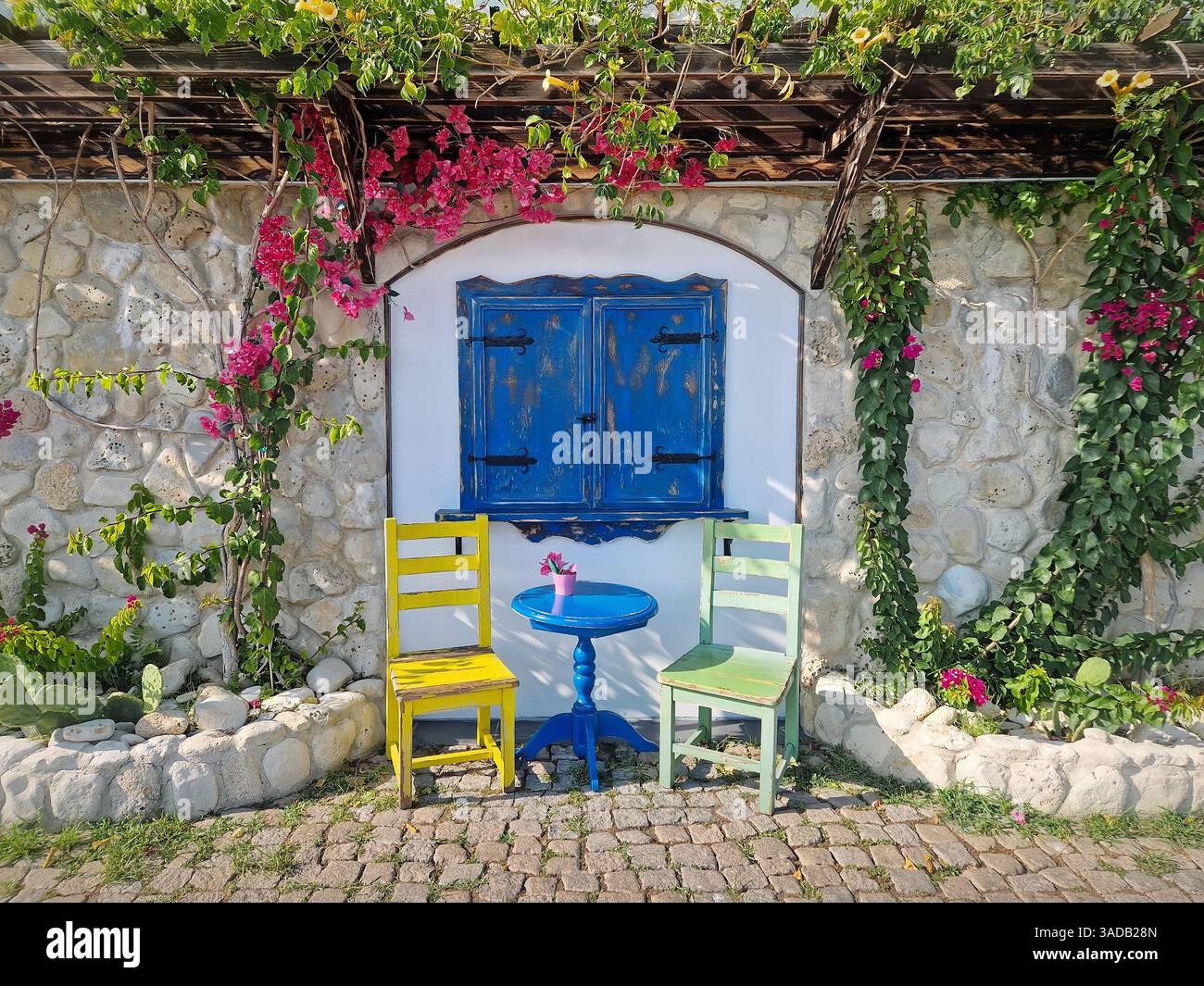 Ästhetisches Café im Hintergrund. Malerische Umgebung im Freien mit einem blauen Holzfenster, zwei farbenfrohen Stühlen und einem kleinen runden Tisch, Steinmauer, verziert - Smartphone-aufgenommenes Stockfoto