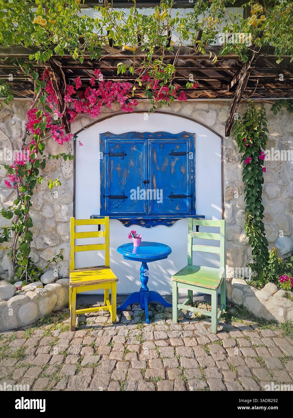 Wunderschöner Hintergrund der Café-Terrasse. Malerische Umgebung im Freien mit einem blauen Holzfenster, zwei farbenfrohen Stühlen und einem kleinen runden Tisch, Steinmauer, verziert - Smartphone-aufgenommenes Stockfoto