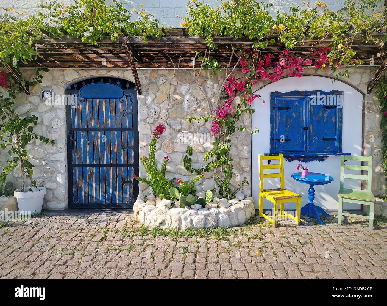 Rustikales mediterranes Café. Steinwände mit leuchtenden Blumen und Grün, blaue Holztür und Fenster, farbenfrohe Stühle und Tische - Smartphone-aufgenommenes Stockfoto