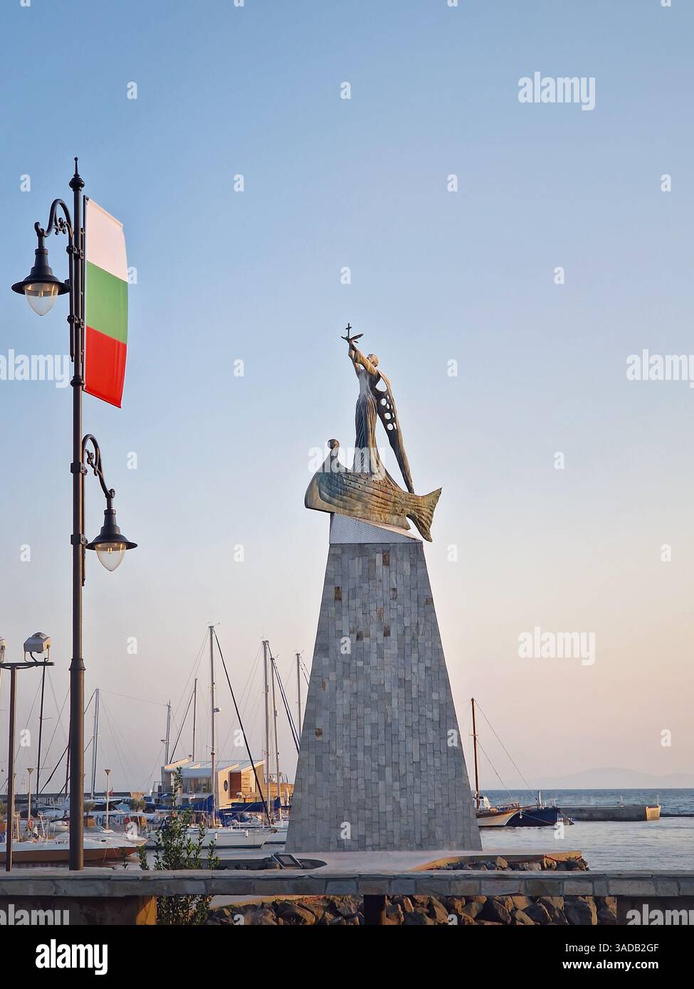 Die Statue des Heiligen Nikolaus Schutzpatron der Seeleute in der Altstadt von Nessebar, Bulgarien. Ruhiger Sonnenuntergang am Hafen mit Booten, die in ruhigem Wasser angedockt sind, ein Stre - Smartphone-aufgenommenes Stockfoto