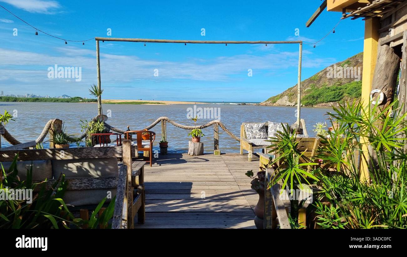 Dieses malerische Foto zeigt eine ruhige Holzterrasse mit Blick auf die Begegnung eines ruhigen Flusses und das offene Meer unter einem leuchtend blauen Himmel. - Smartphone-aufgenommenes Stockfoto