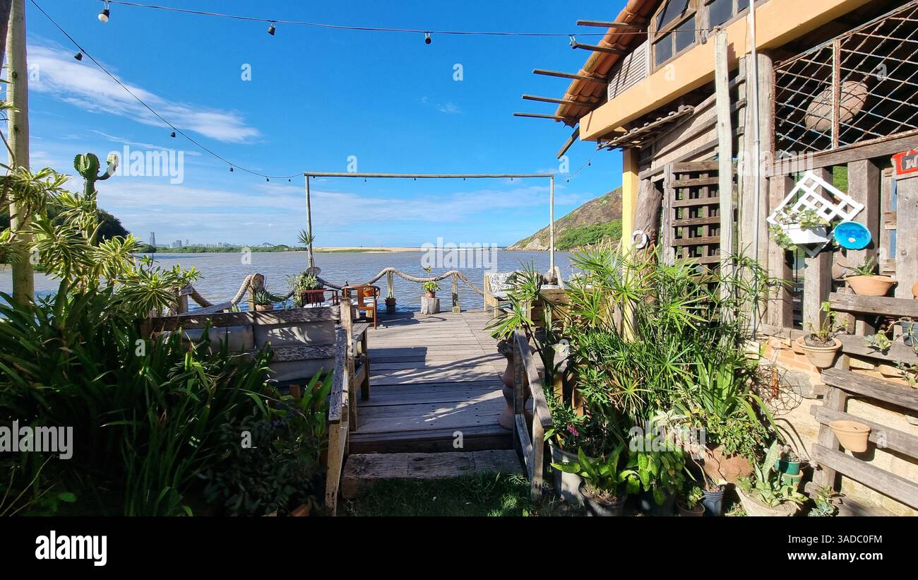Dieses malerische Foto zeigt eine ruhige Holzterrasse mit Blick auf die Begegnung eines ruhigen Flusses und das offene Meer unter einem leuchtend blauen Himmel. - Smartphone-aufgenommenes Stockfoto