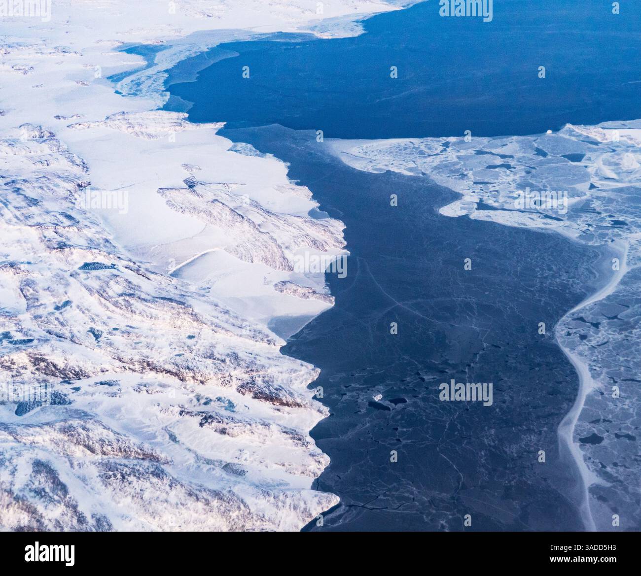 Luftaufnahme einer gefrorenen Landschaft mit schneebedecktem Land, das auf ein teilweise gefrorenes Gewässer trifft. Stockfoto