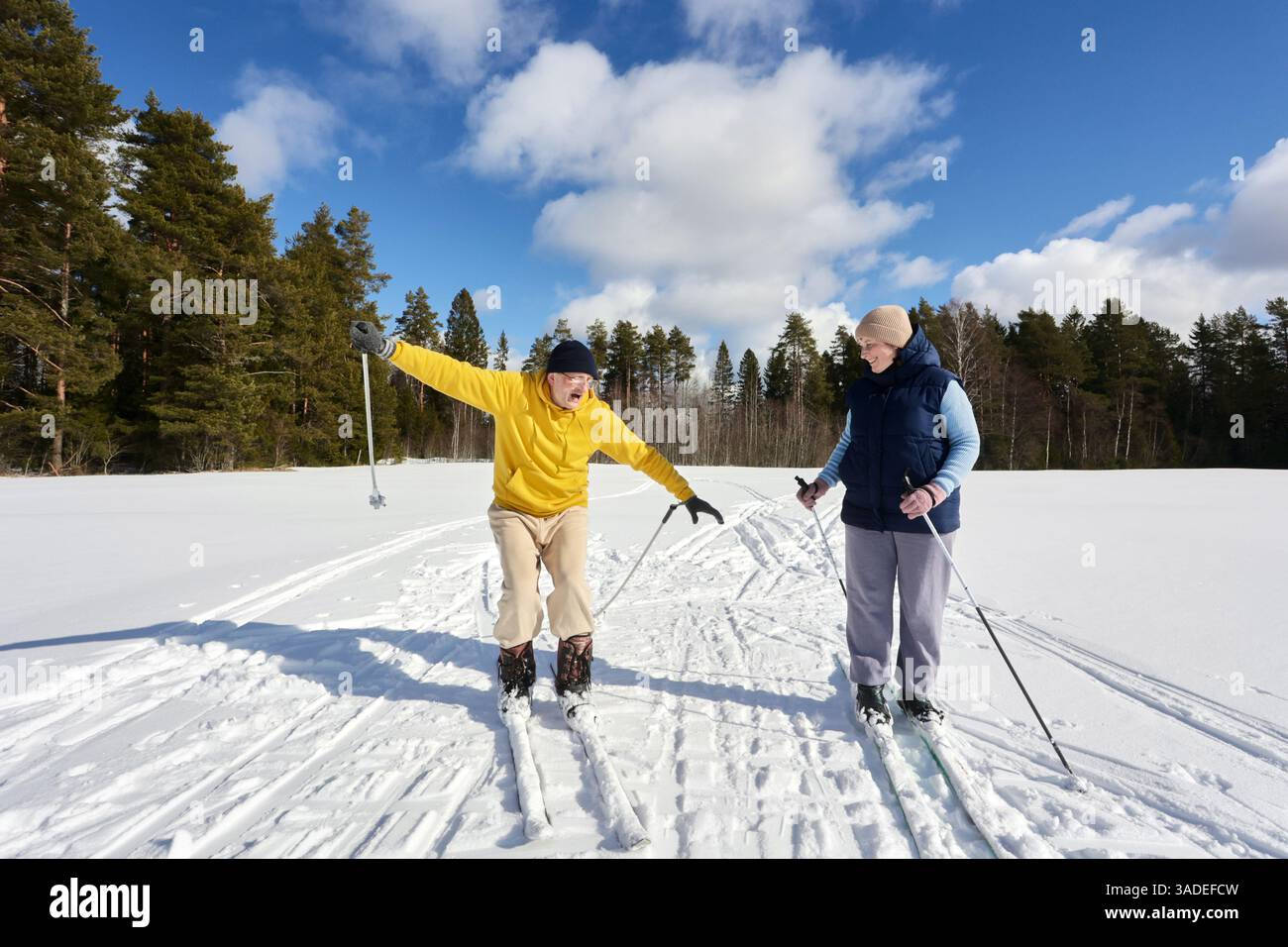 Ein älteres erwachsenes Paar fährt Ski, der Mann stolpert und wirft überraschend die Arme aus, während die Frau sanft lächelt, helle Winterszene mit Waldkulisse. Stockfoto