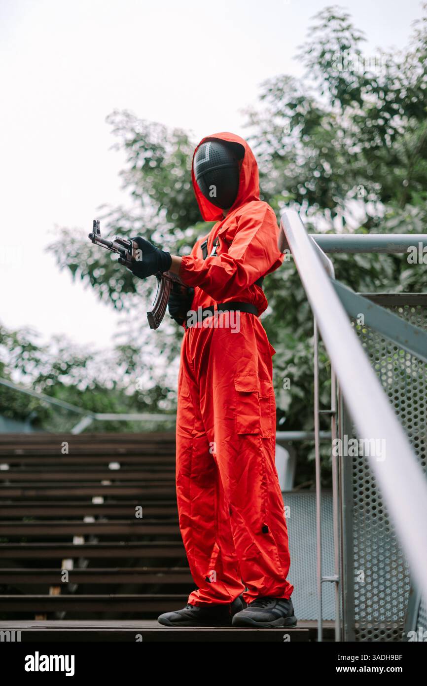 Jakarta, Indonesien, 4. April 2025. Der maskierte Mann in rotem Jumpsuit steht auf der Treppe und hält eine Waffe und erschafft ein mysteriöses und leicht beunruhigendes atmos Stockfoto