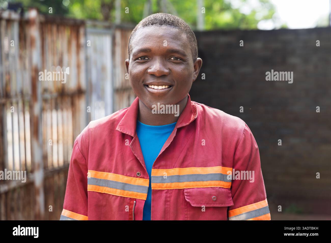 Porträt eines einzelnen afrikanischen Dorfes glücklicher lächelnder Arbeiter Mann in Arbeitskleidung, der im Hof steht, junger Mann Stockfoto