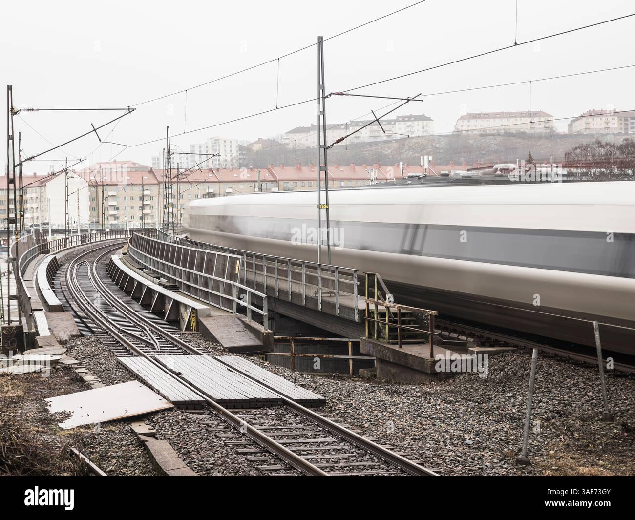 In Göteborg, Schweden, rastet ein schlanker Zug auf kurvenreichen Strecken vorbei. Graue Himmel und Nebel schaffen eine einzigartige Atmosphäre, da Gebäude ruhig in der Nähe stehen, Hi Stockfoto