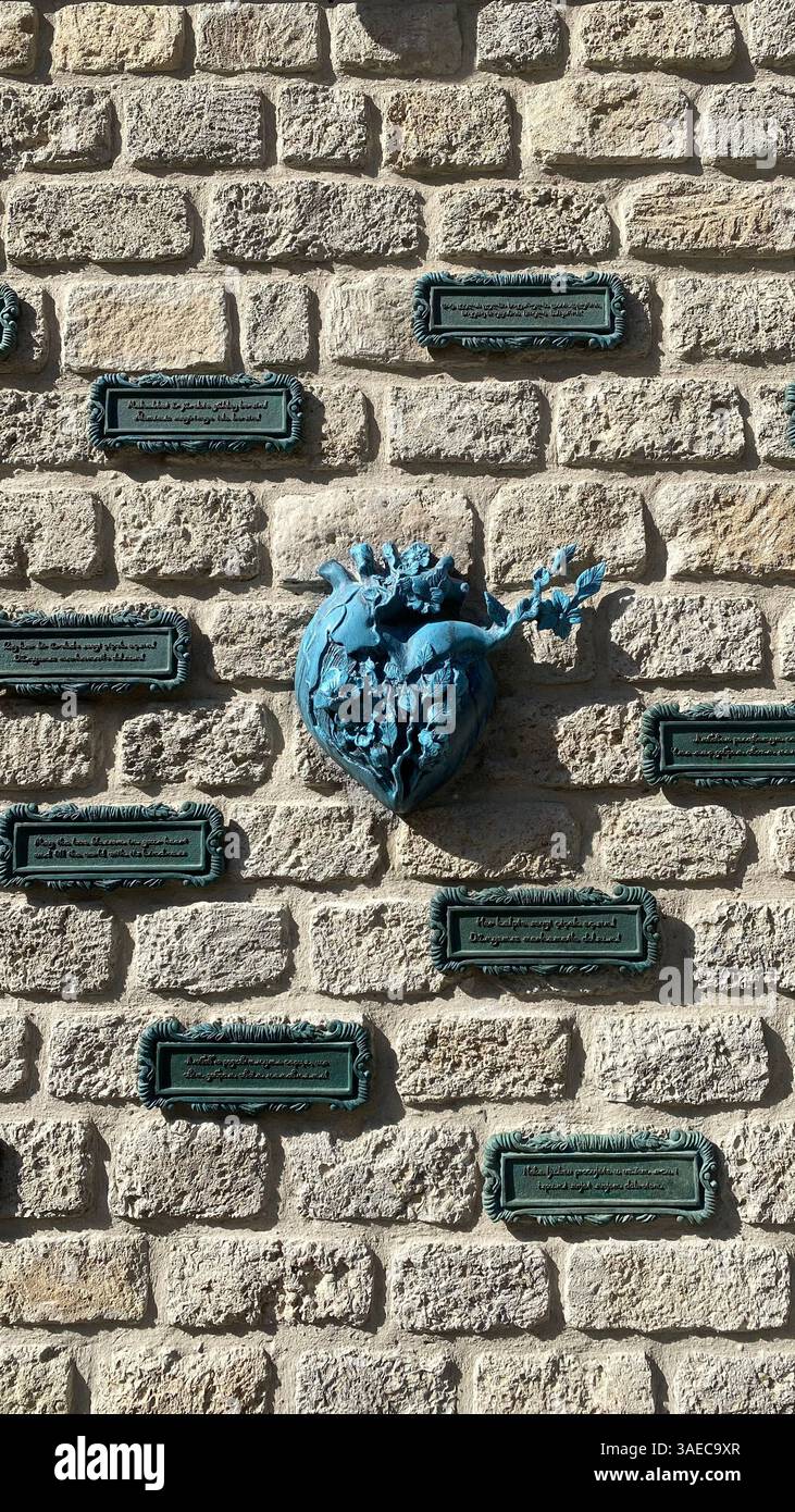 Ein Herz mit floralen Details ist auf einer strukturierten Steinwand montiert, umgeben von gerahmten Tafeln mit mehrsprachigen Inschriften. Urbane Kunst trifft auf - Smartphone-aufgenommenes Stockfoto