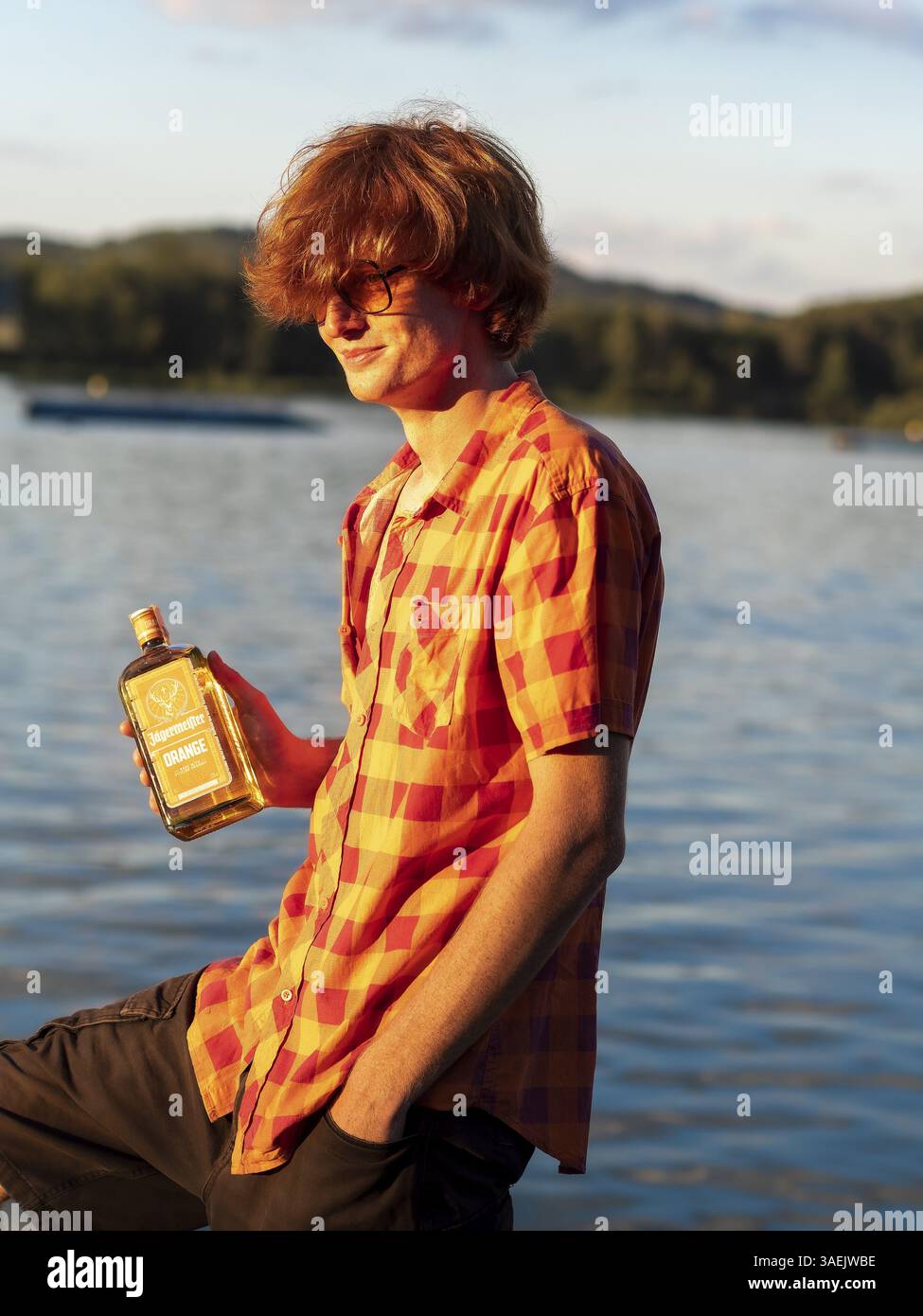 Junger Mann, der am See steht, eine Flasche Alkohol in den Händen hält, Kräuterlikör, rote Haare, orangenes Hemd Stockfoto