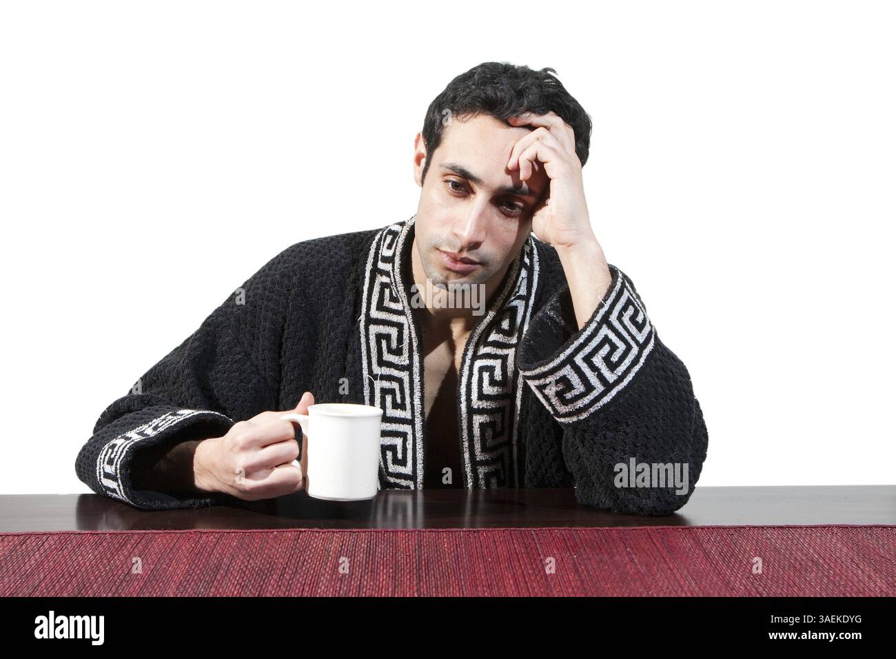 Gutaussehender Kerl am Morgen, der gerade an einem Tisch in seinem Gewand aufwachte, mit einer Tasse, die sich von seinem Kater erholte, isoliert auf White, New York, USA Stockfoto