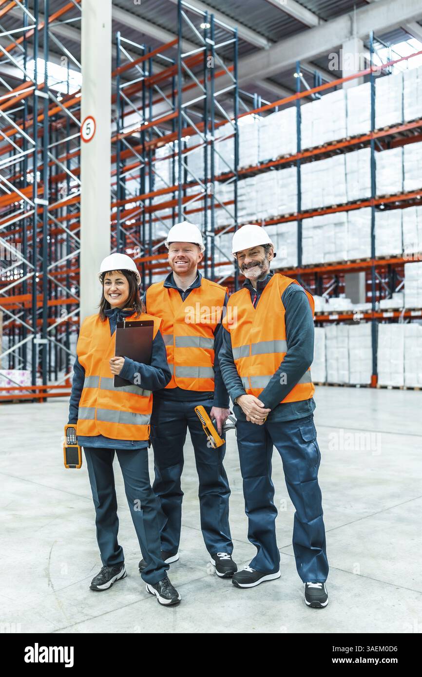 Lagerarbeiter, die Sicherheitswesten und Helme tragen, lächeln, während sie in einem großen Lager Werkzeuge zur Bestandsverwaltung halten Stockfoto