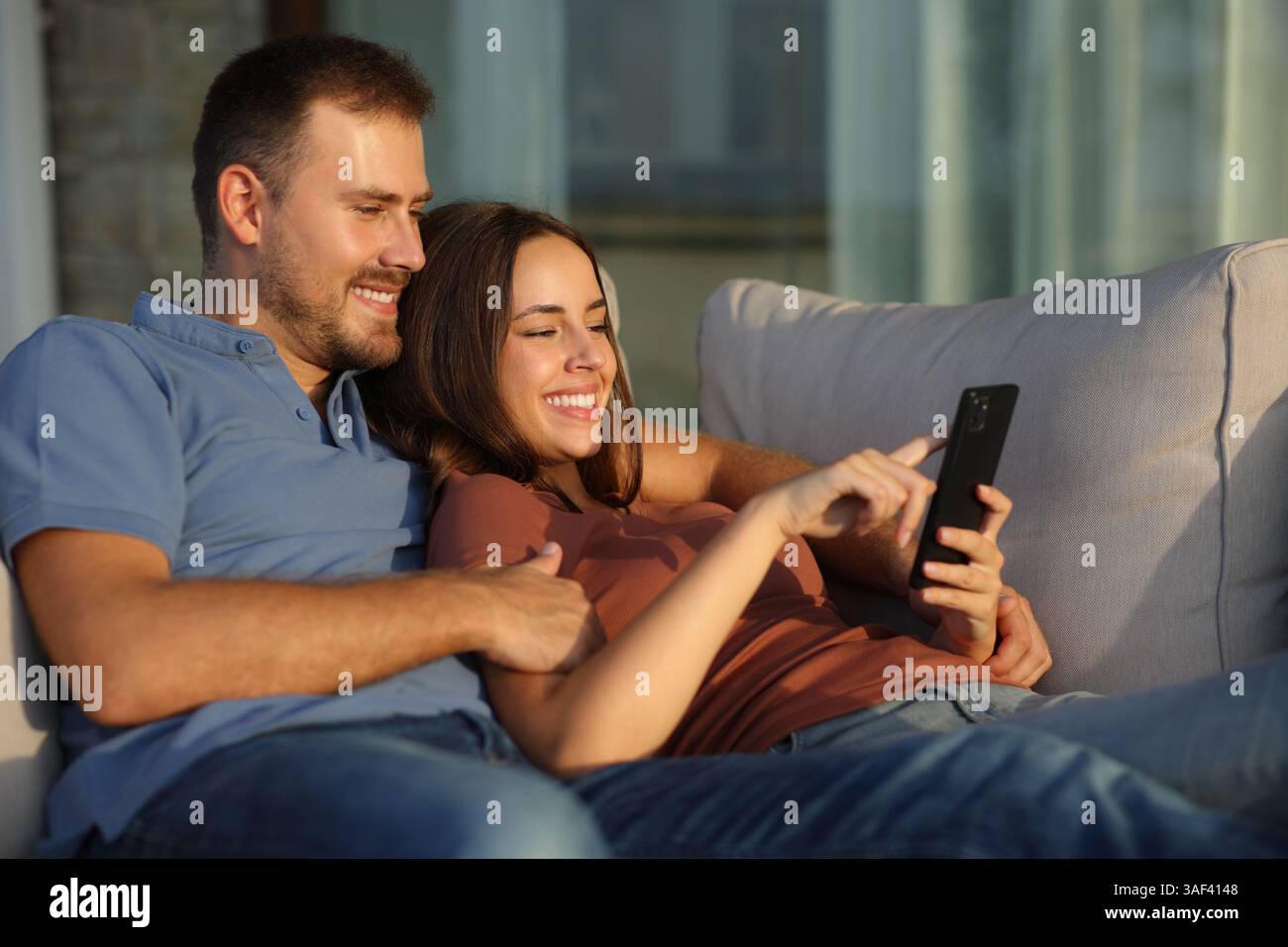 Glückliches Paar, das das Handy benutzt und auf einem Sofa auf einer Terrasse sitzt, zu Hause oder im Hotel bei Sonnenuntergang Stockfoto