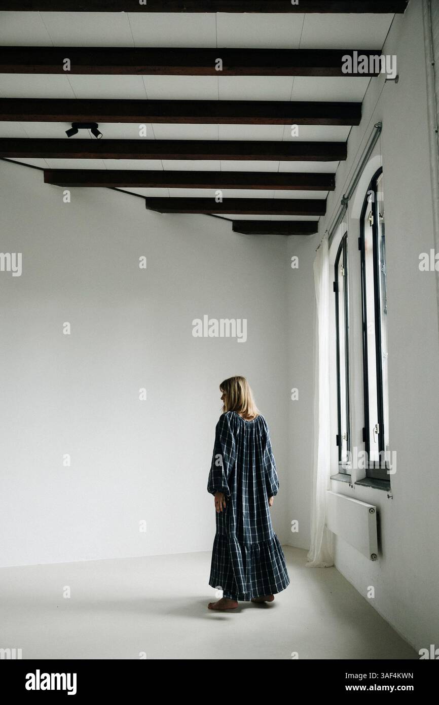 Frau in kariertem Kleid barfuß in der Nähe eines hellen Fensters in einem minimalen Raum Stockfoto