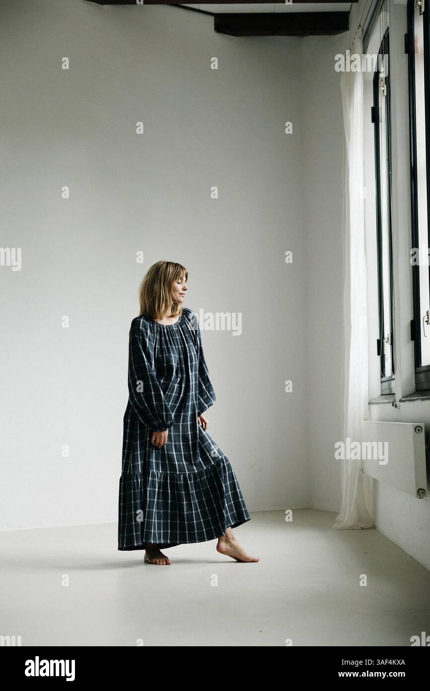 Frau in kariertem Kleid barfuß in der Nähe eines hellen Fensters in einem minimalen Raum Stockfoto