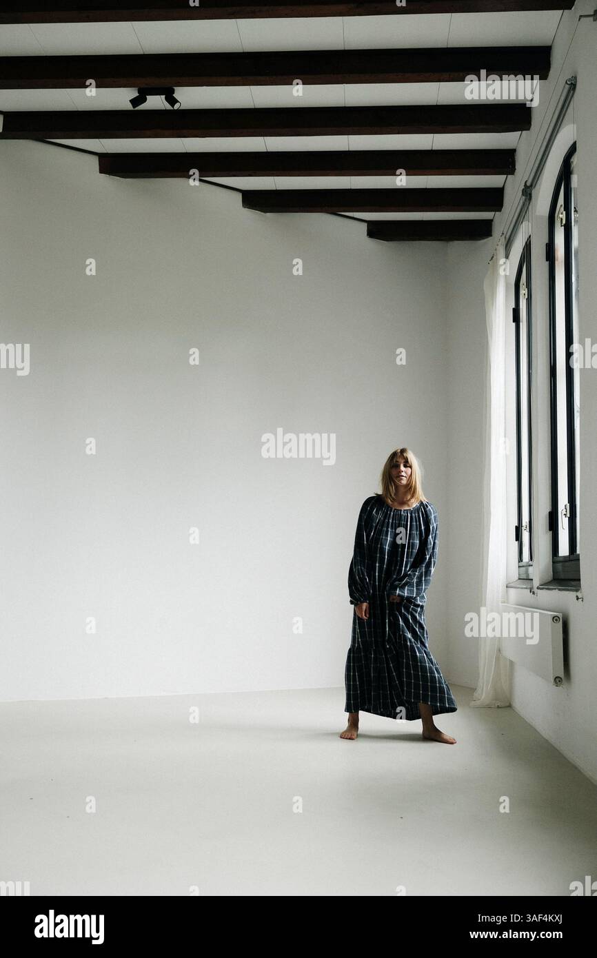 Frau in kariertem Kleid barfuß in der Nähe eines hellen Fensters in einem minimalen Raum Stockfoto