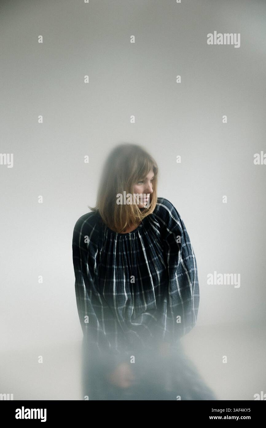 Frau in kariertem Kleid barfuß in der Nähe eines hellen Fensters in einem minimalen Raum Stockfoto