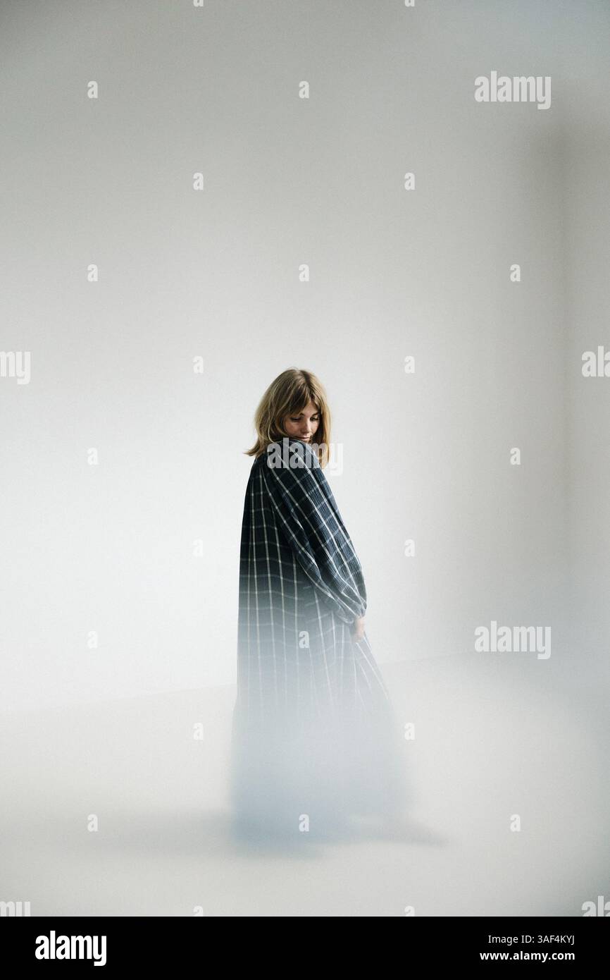 Frau in kariertem Kleid barfuß in der Nähe eines hellen Fensters in einem minimalen Raum Stockfoto