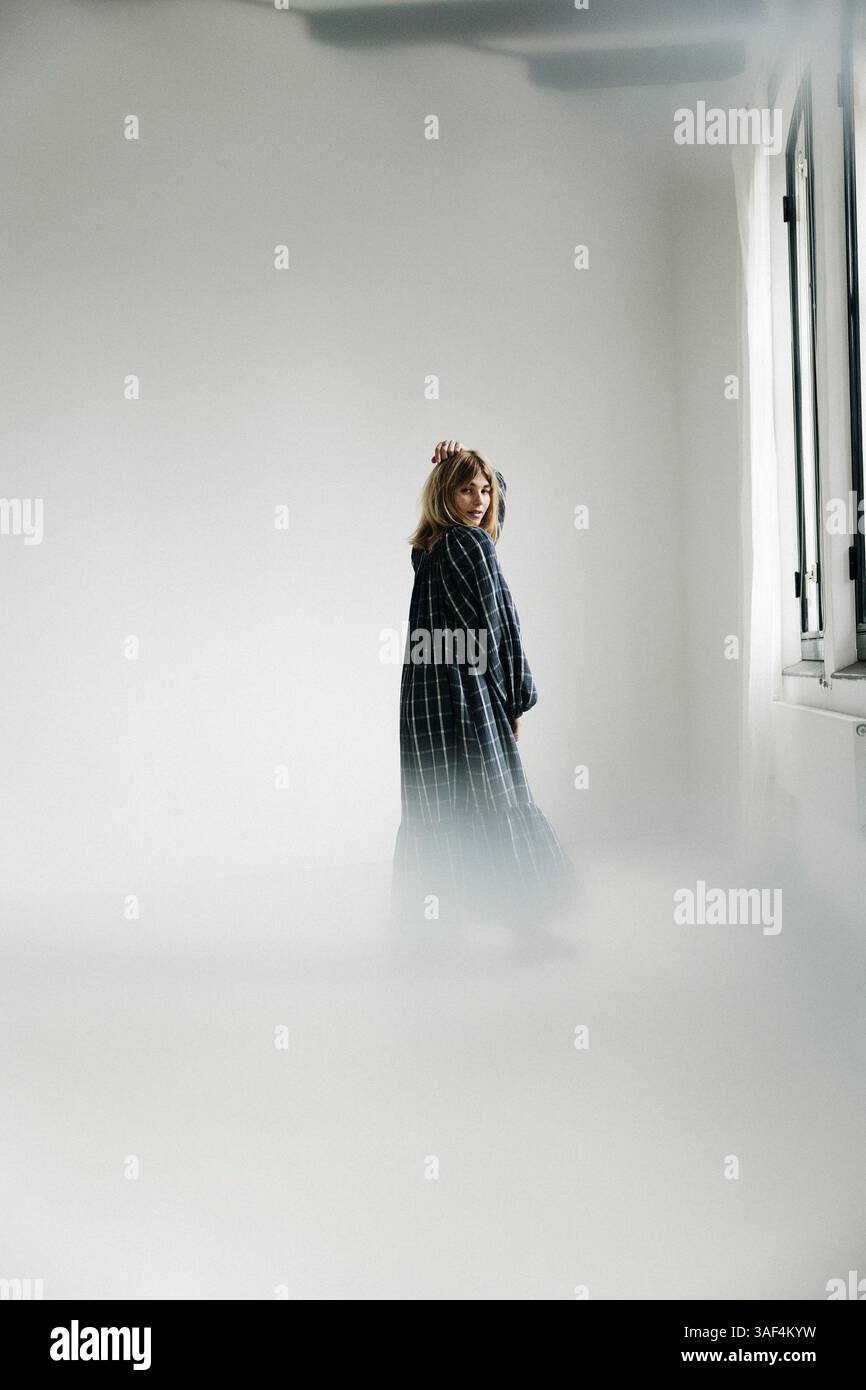 Frau in kariertem Kleid barfuß in der Nähe eines hellen Fensters in einem minimalen Raum Stockfoto