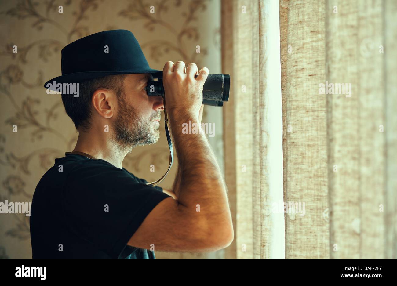 Mann mit Fedora-Hut, der durch ein Fernglas schaut und vom Fenster aus ausspioniert. Spion- und Suchkonzept Stockfoto