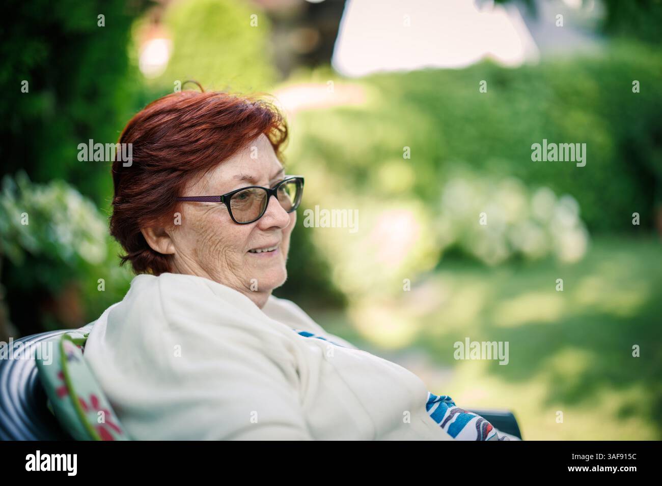 Lächelnde Seniorin mit rotem Haar und Brille, die im Garten sitzt und ihren Ruhestand und ihre goldenen Jahre genießt. Ehrliches Porträt Stockfoto