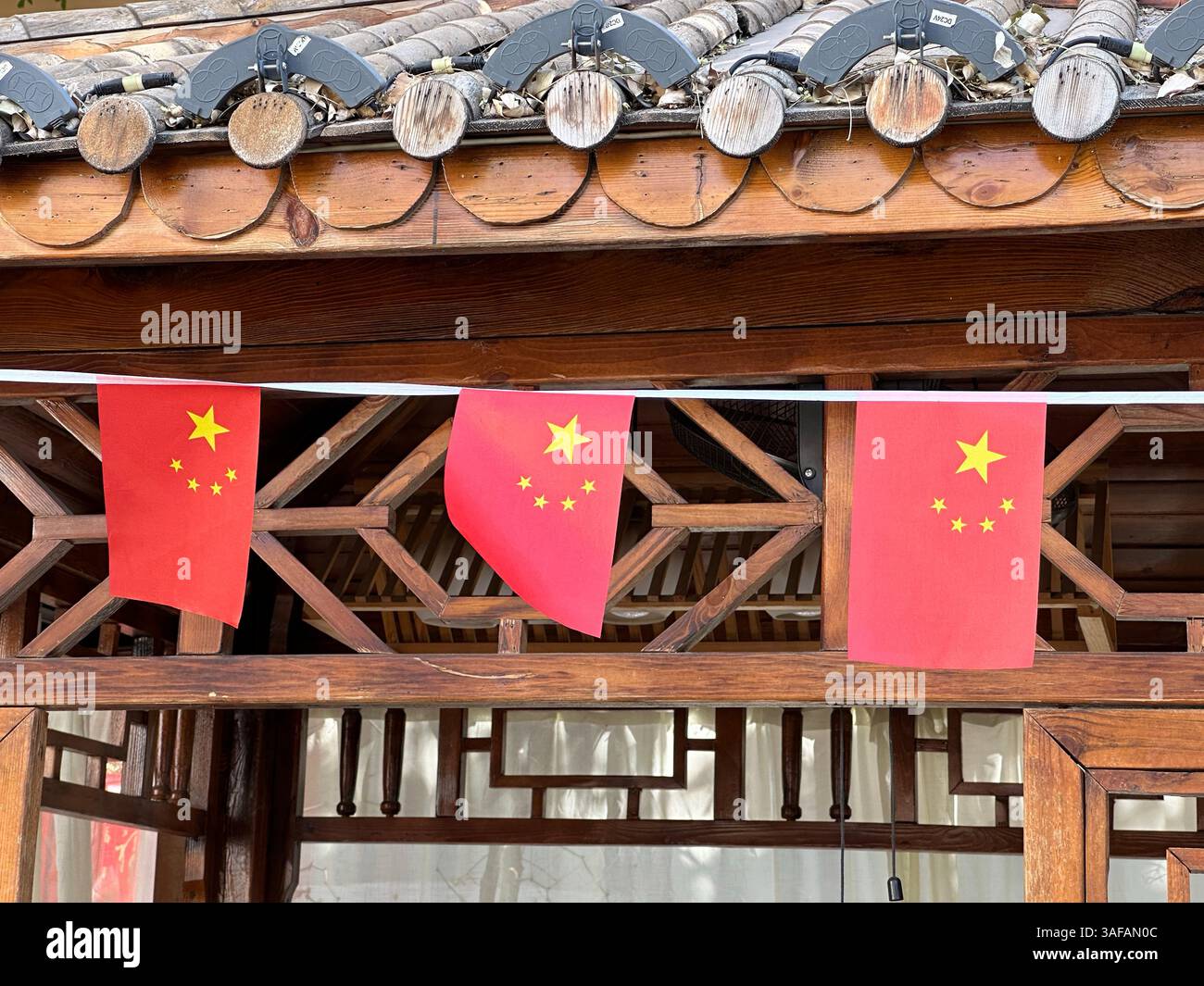 Chinesische Flaggen mit Sternenmotiv, die an einer traditionellen Holzstruktur hängen, kulturell. - Smartphone-aufgenommenes Stockfoto
