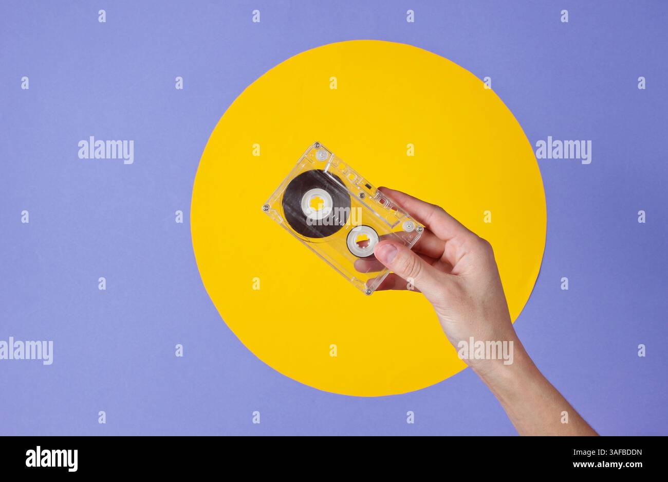 Weibliche Hand, die Audiokassette auf violettem Hintergrund mit gelbem Kreis hält Stockfoto