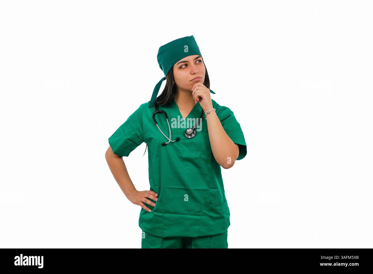 Nachdenkliche lateinische Chirurgin, die grüne medizinische Peelings und Stethoskop trägt, denkt mit der Hand auf ihrem Kinn und blickt auf einem weißen Hintergrund weg Stockfoto