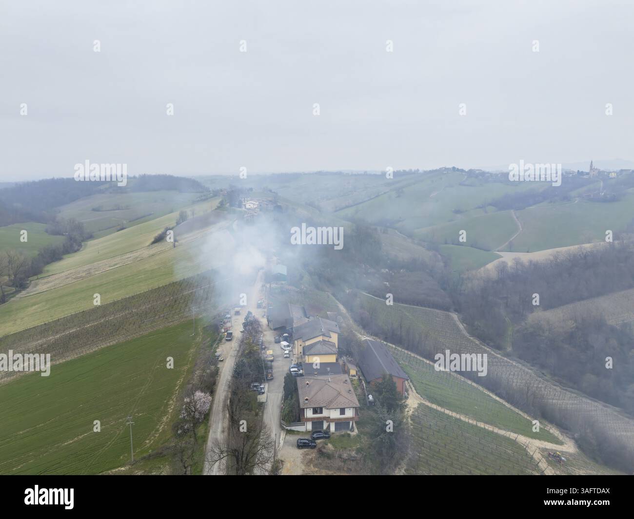 Rauch steigt aus brennendem Ackerschnitt in einem Weinberg auf, mit einem Weingut im Hintergrund, in der wunderschönen Landschaft der Martani-Hügel in Piacenza, I Stockfoto