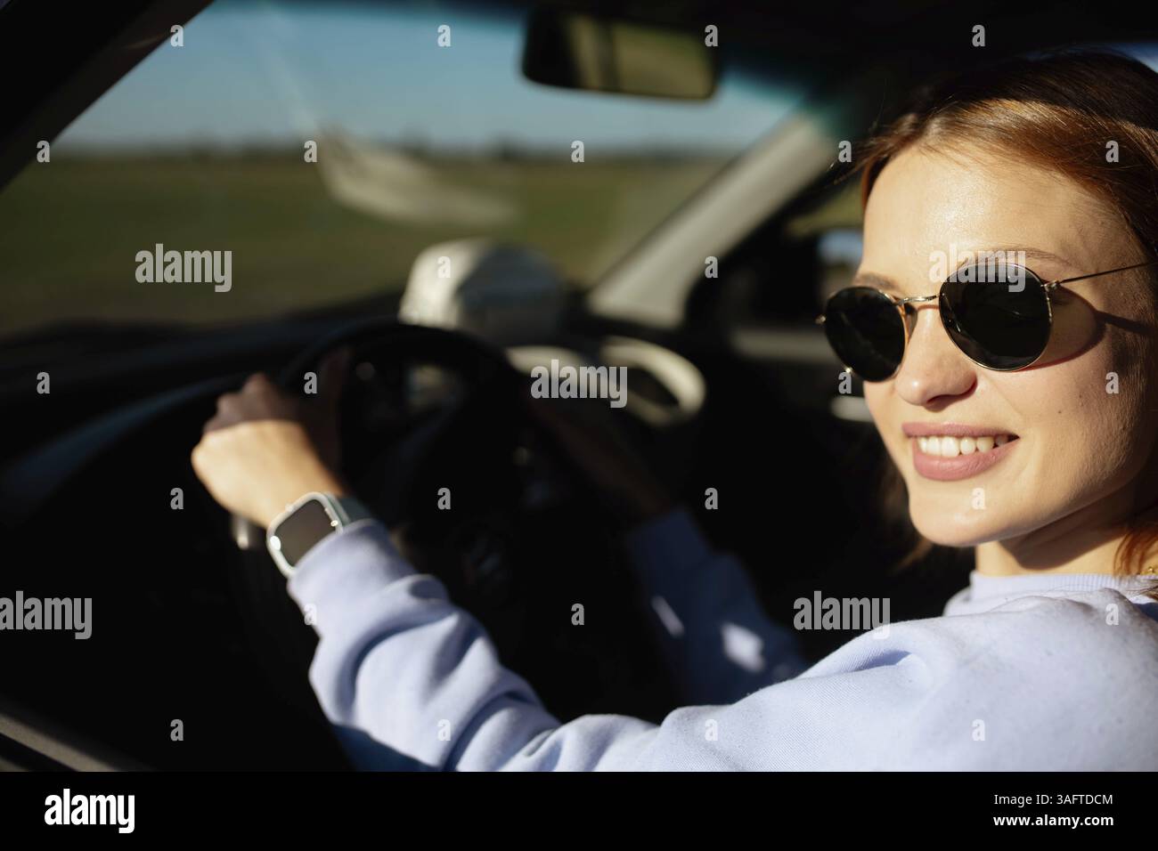 Frau lächelt und fährt ein Auto mit Sonnenbrille in hellem Sonnenlicht Stockfoto