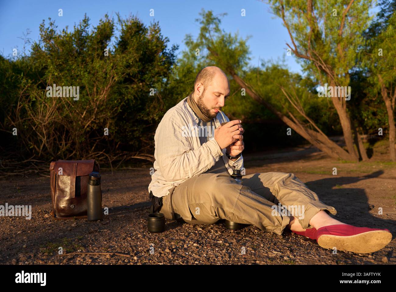 Hispanischer Reisender, der bei Sonnenaufgang am Ufer eines Baches sitzt und die ersten Sonnenstrahlen empfängt, während er Mate trinkt, ein typisches argentinisches Getränk. El Stockfoto