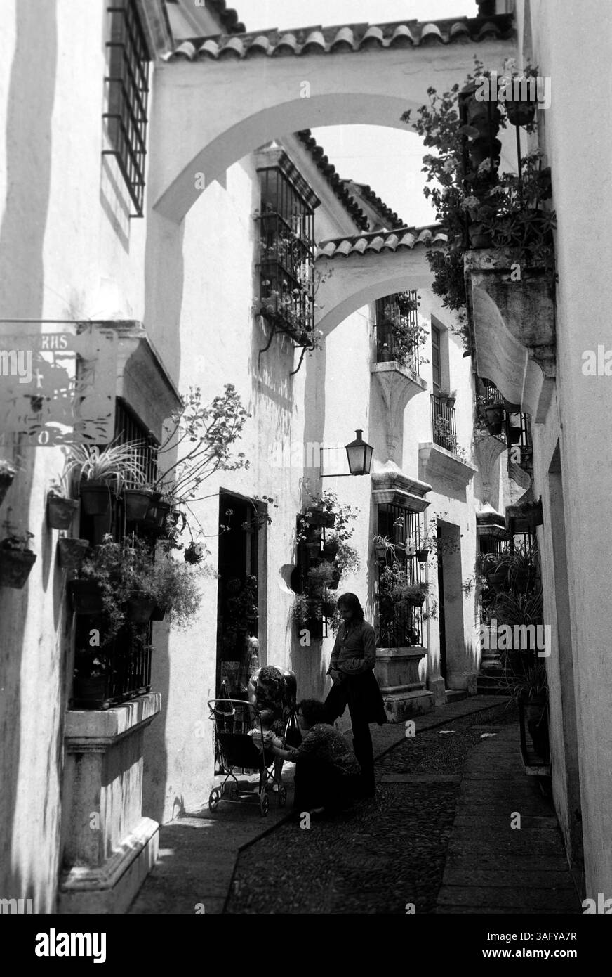 In einer engen Gasse zwischen weißgetünchten, zweistöckigen Häusern mit Topfpflanzen in den vergitterten Fenstern kümmert sich zwei Frauen um ein Kleinkind in einem Kinderwagen, ein junger Mann neben ihnen sieht dabei zu, Costa Brava, 1975. In einer engen Gasse zwischen weiß getünchten, zweistöckigen Häusern mit Topfpflanzen in den vergitterten Fenstern kümmern sich zwei Frauen um ein Kleinkind in einem Kinderwagen, ein junger Mann neben ihnen schaut zu, Costa Brava, 1975. Stockfoto