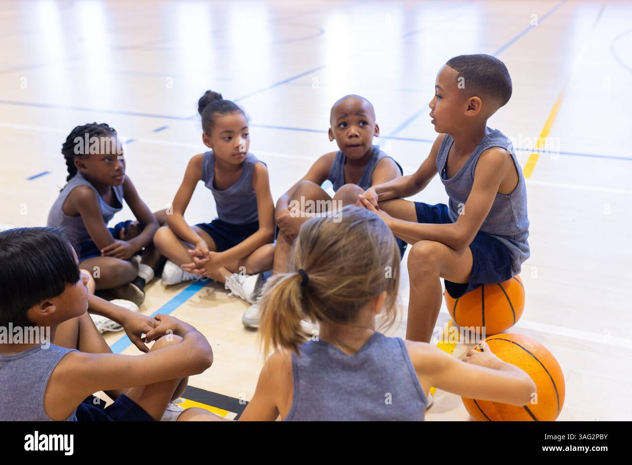 Im Schulstudio sitzen multirassische Kinder auf dem Boden und lauschen dem Teamkollegen, der Basketball hält Stockfoto