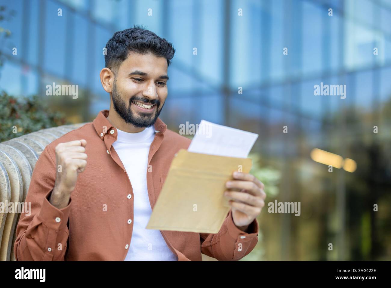 Ein junger Mann feiert mit Aufregung und enthüllt ein positives Briefergebnis mit geballter Faust und einem großen Lächeln im Freien. Stockfoto