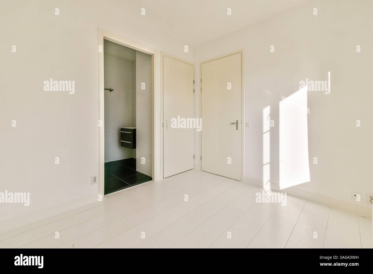 Ein helles und geräumiges Interieur mit weißen Wänden, hellem Holzfußboden und natürlichem Licht, das Schatten wirft. Eine offene Tür führt zu einem anderen Zimmer. Stockfoto