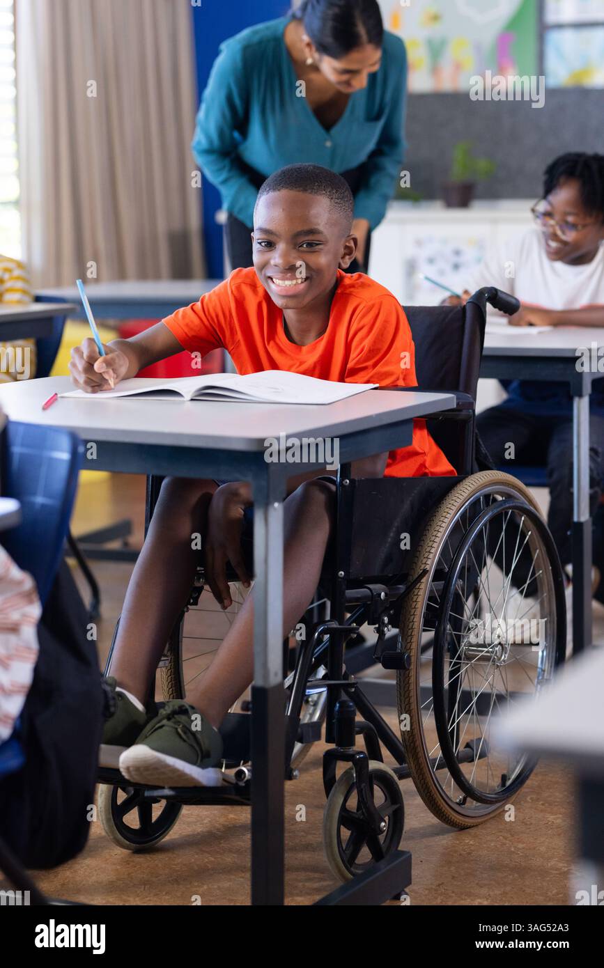 In der Schule, Junge im Rollstuhl, schrieb in Notizbuch, lächelte im Klassenzimmer Stockfoto