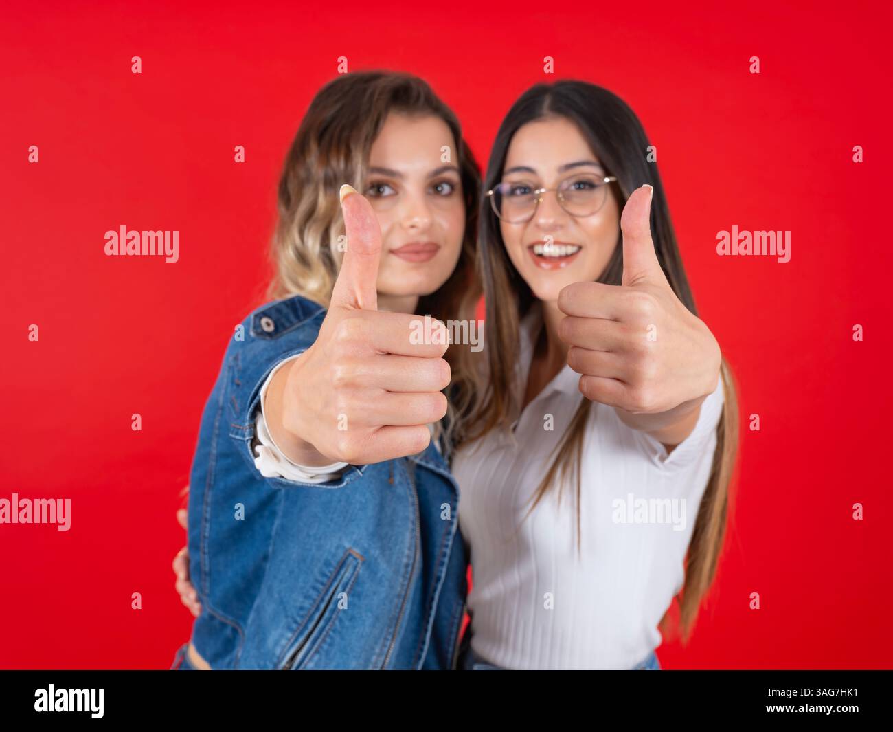 Charmante zwei kaukasische, selbstbewusste Frauen, die zusammen den Daumen hoch über rotem Studiohintergrund zeigen. Selektiver Fokus auf Hands Approve, Offer Concept Image Stockfoto