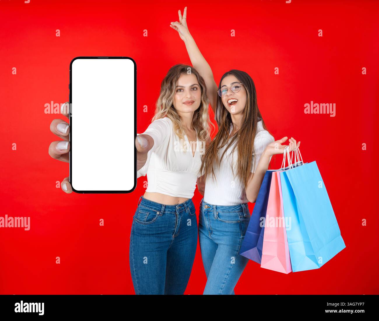 Zwei glückliche Käuferinnen, die über rotem Hintergrund stehen und eine halten, die ein leeres Handy zeigt, verspotten eine mit Einkaufstaschen Stockfoto
