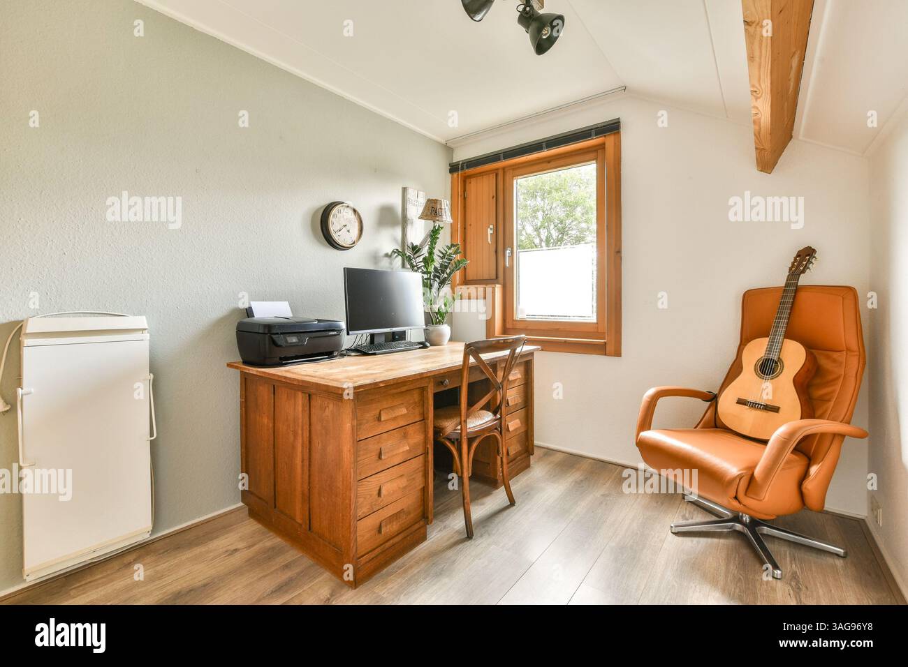 Ein stilvolles Home Office mit Holzschreibtisch, Computer-Setup und einladendem Sessel für eine ausgewogene und produktive Arbeitsumgebung. Stockfoto