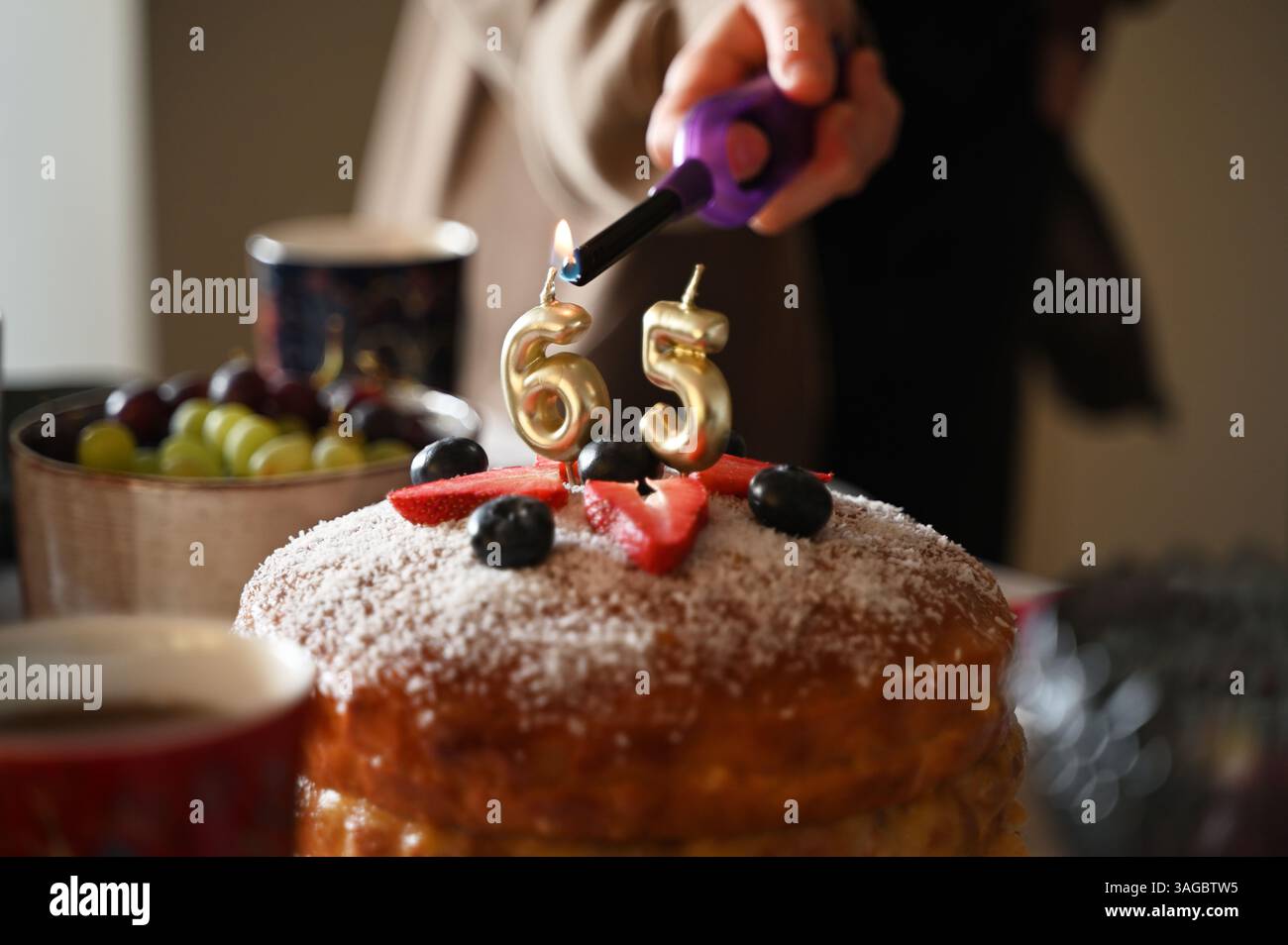 Ein festliches Nahaufnahme-Bild, das den Moment festmacht, in dem Kerzen auf einer Geburtstagskuche zum 65. Geburtstag angezündet werden. Der Kuchen ist mit frischen s dekoriert Stockfoto