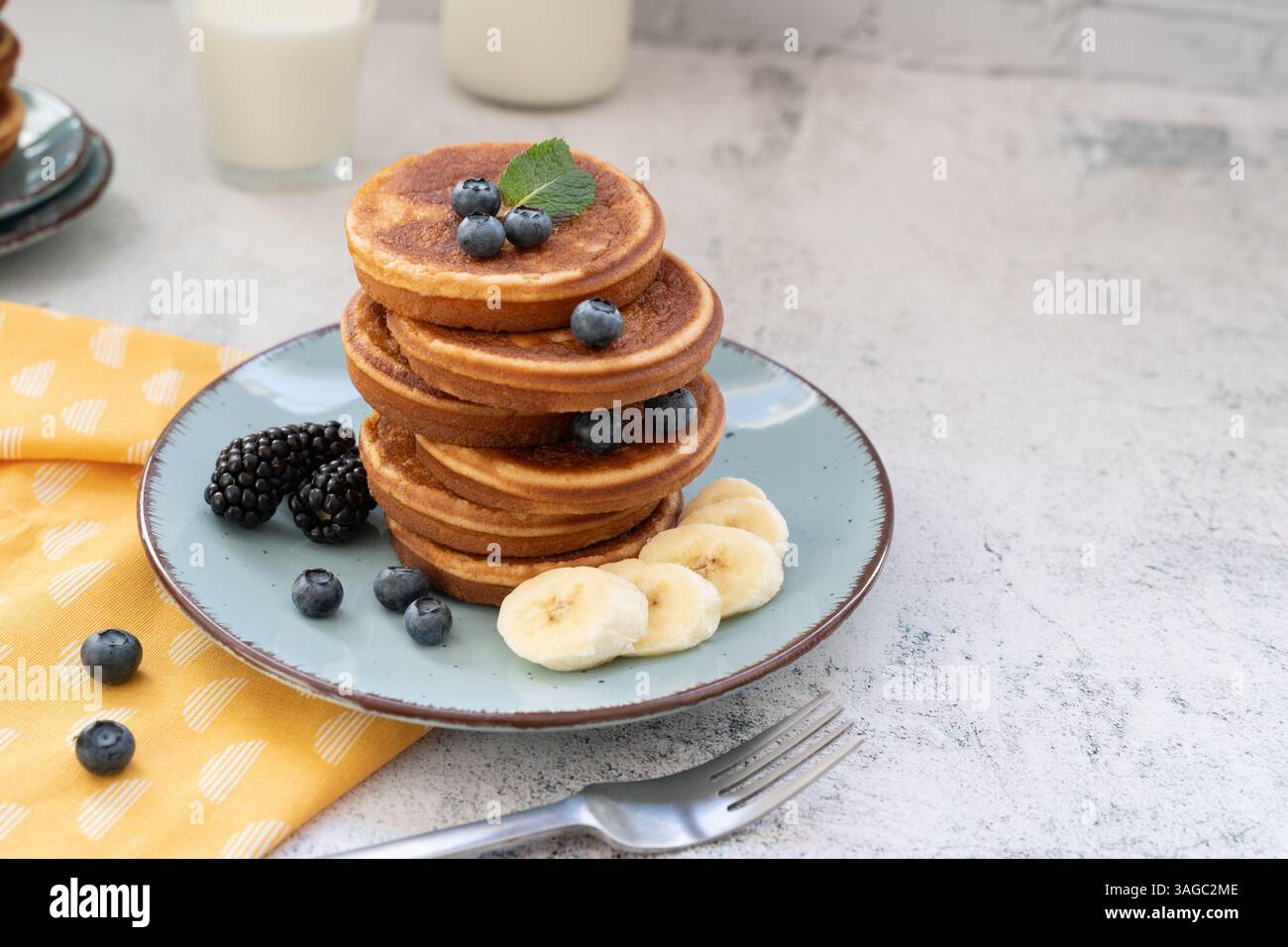 Süße und flauschige, gestapelte Pfannkuchen mit Bananen und Beeren. Mit Honig auf blauen Tellern gegen hellgraue Tischplatte mit gelbem Tuch Stockfoto