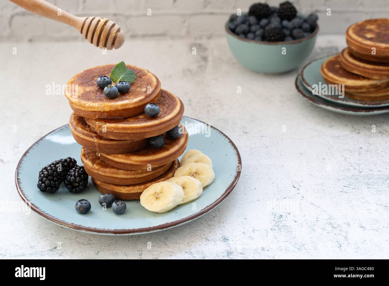 Zwei Stapel süßer und flauschiger Pfannkuchen mit Bananen und Beeren, auf blauen Tellern vor hellgrauer Tischplatte mit gelbem Tuch Stockfoto