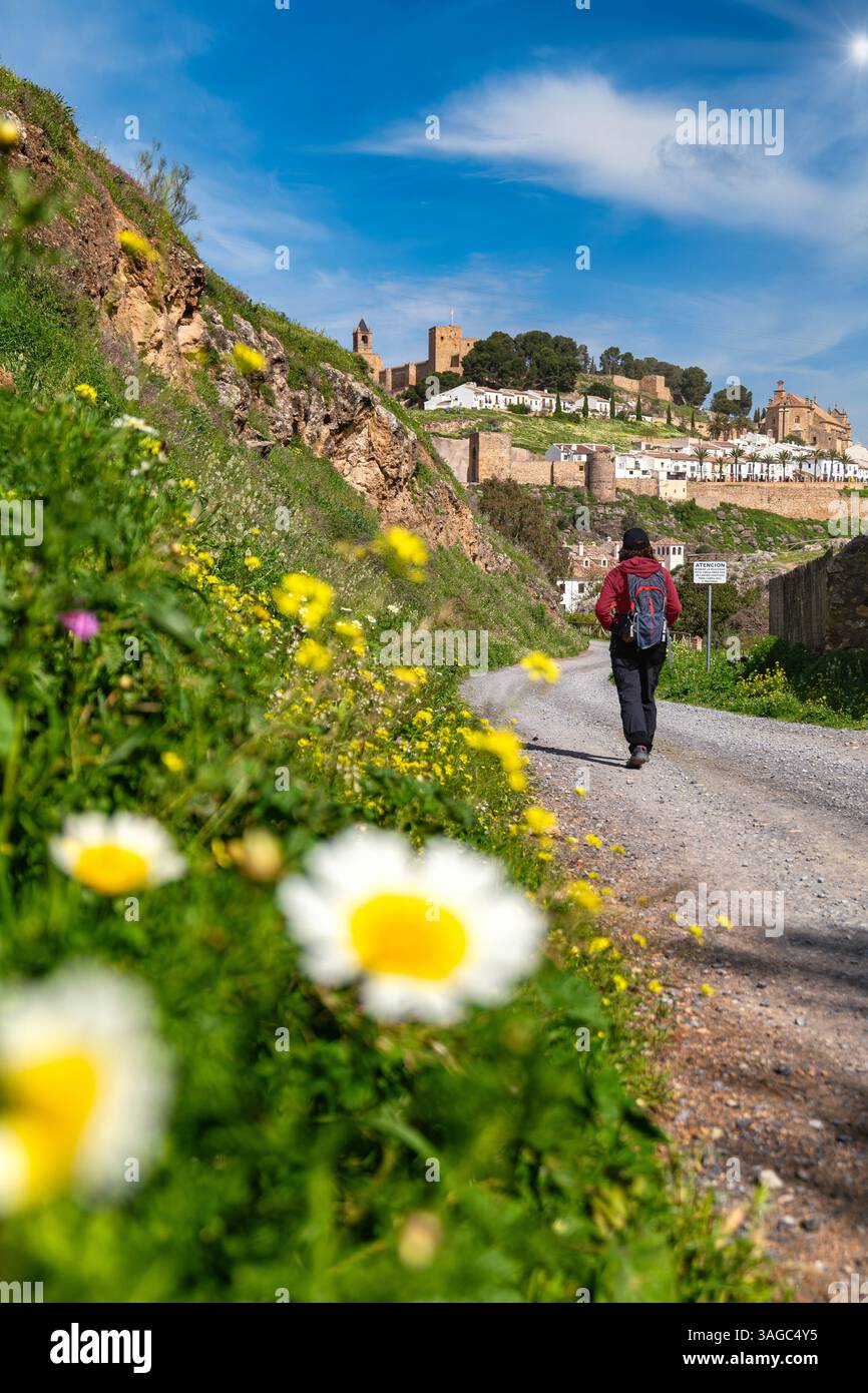 Frau auf dem Rücken, die mit Rucksack auf einem Landweg in Richtung eines Dorfes mit Schloss geht. Im Vordergrund unscharf. Langsamer Tourismus, ländlich Stockfoto