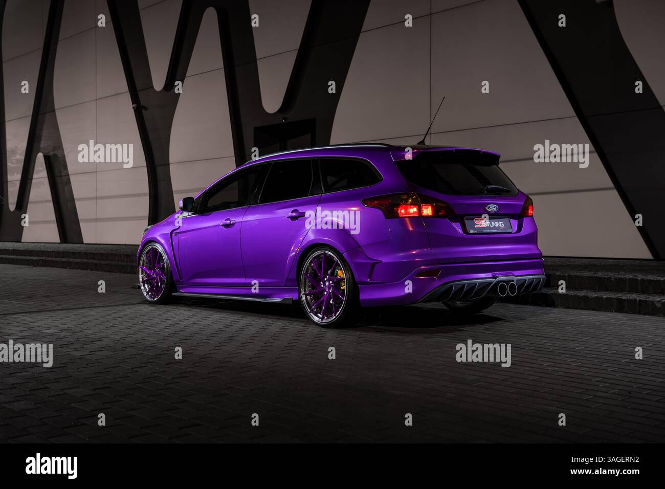 Violettes Ford Focus mk3 parkte nachts in der Nähe eines modernen beigefarbenen Gebäudes. Dreiviertelansicht des sportlichen Focus ST-Wagens hinten. Stockfoto