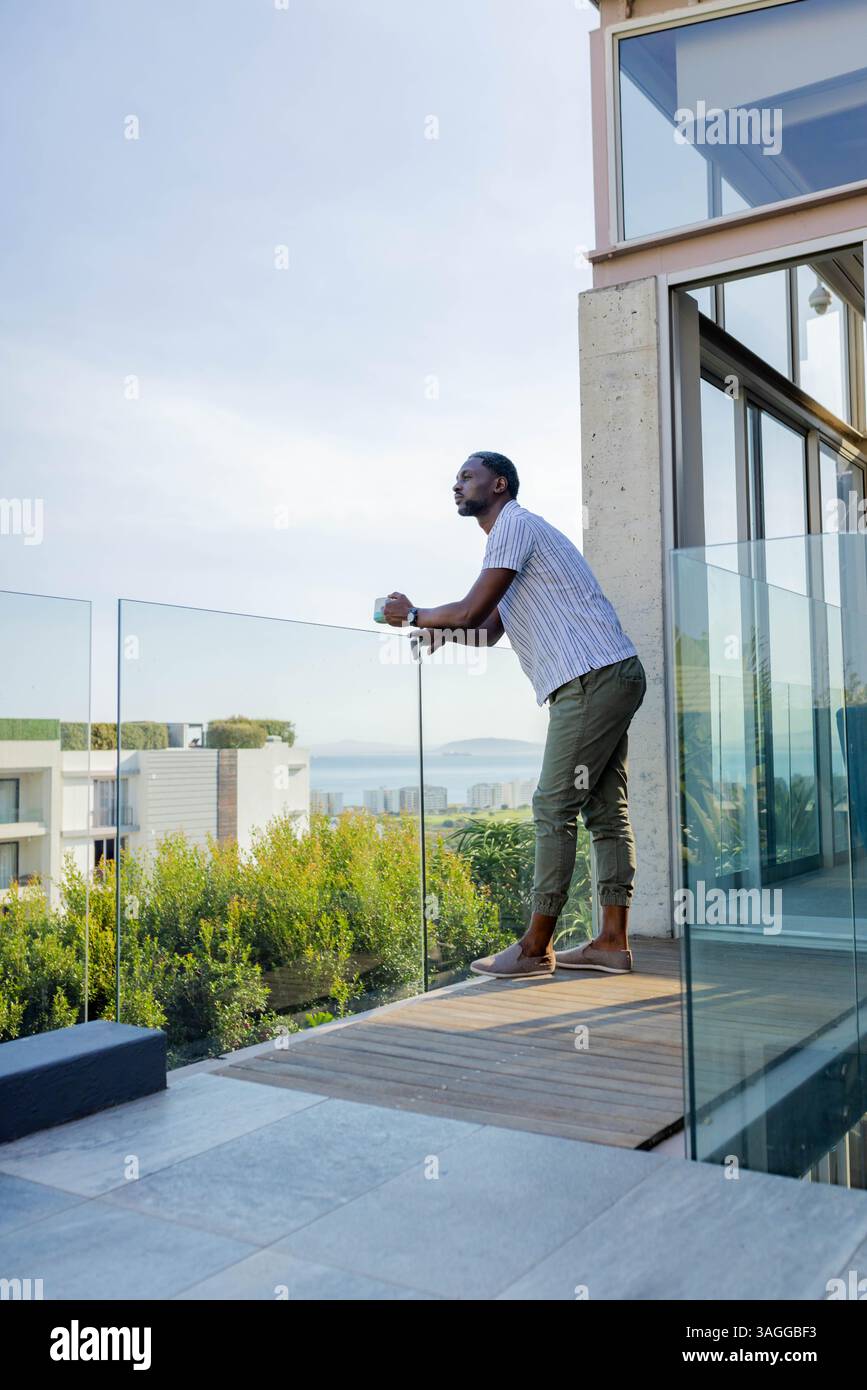 Ein afroamerikanischer Mann auf dem Balkon trinkt morgens Kaffee und blickt auf eine ruhige Landschaft und fühlt sich entspannt. Entspannung, morgendliche Routine, Ruhe, Frieden Stockfoto