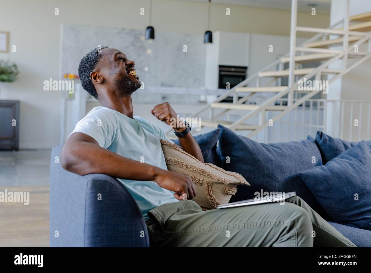 Wir feiern den Erfolg, Afroamerikaner sitzt auf Sofa mit Laptop zu Hause. Leistung, Glück, Arbeit, Home Office, freudig, Stockfoto