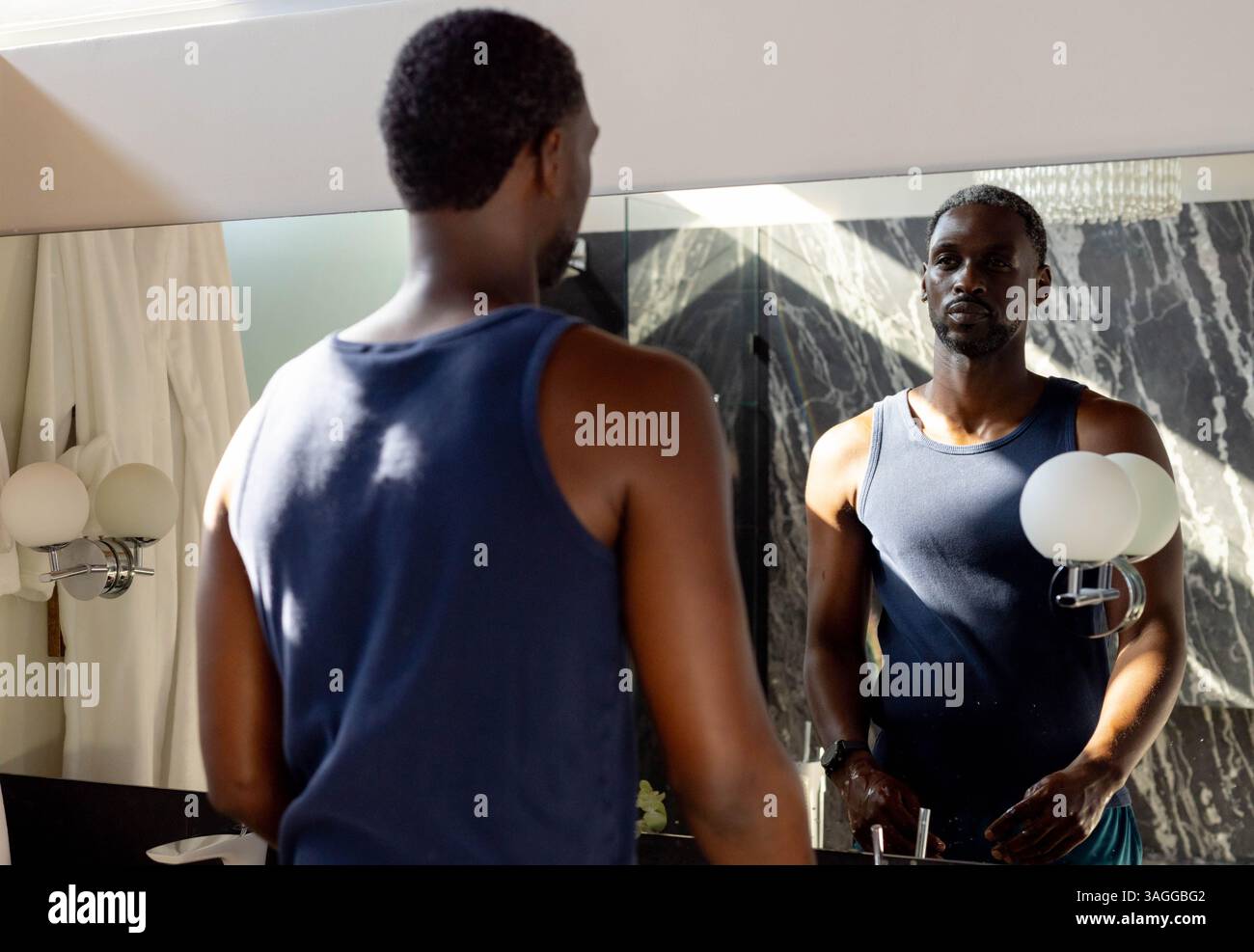 Afroamerikaner im Badezimmer, der auf den Spiegel blickt, Tanktop trägt und nachdenklich reflektiert. Reflexion, Besinnung, Einsamkeit, persönlich, Pflege Stockfoto