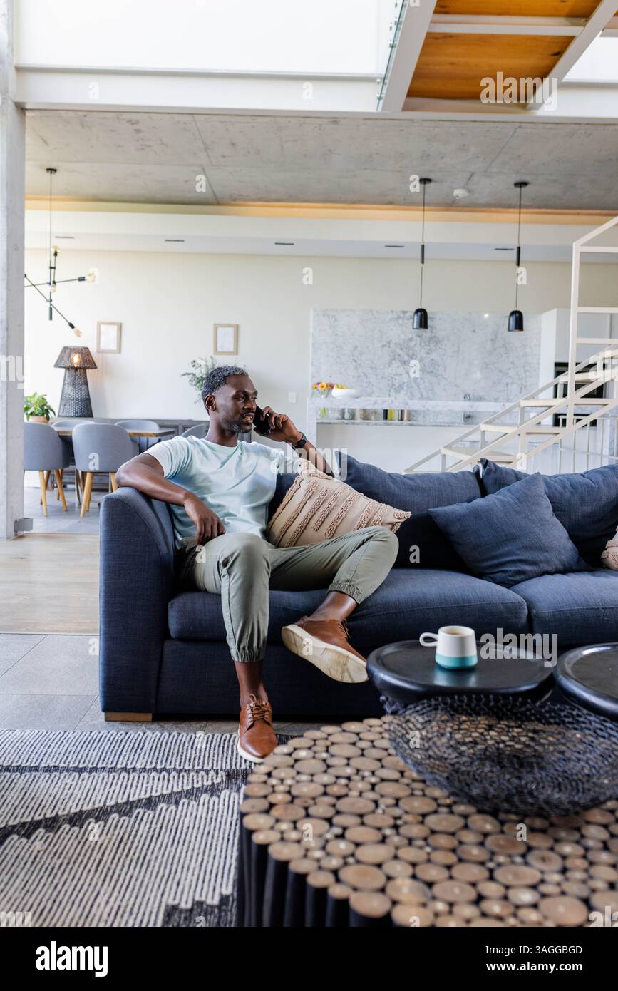 Afroamerikaner sitzt zu Hause auf dem Sofa, spricht auf dem Smartphone, sieht entspannt aus. Konversation, Kommunikation, Technologie, Entspannung, Haushalt, l Stockfoto