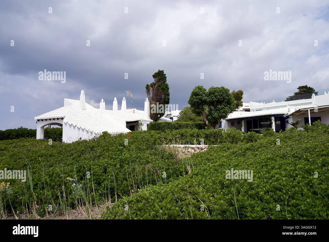 Ein malerischer Blick auf Binibeca, Menorca, mit weißen Gebäuden vor einem Hintergrund von üppigem Grün unter einem bewölkten Himmel. Stockfoto
